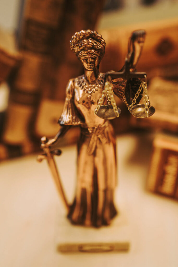 Bronzene Justitia-Statue mit Schwert und Waage, unscharfe Bücher im Hintergrund.