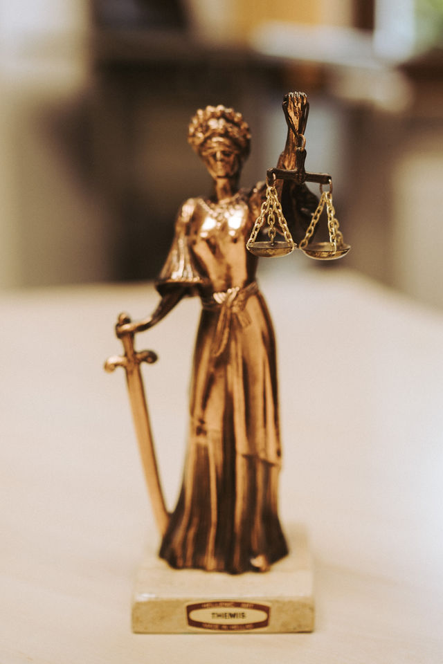 Bronzene Justitia-Statue mit Schwert und Waage, unscharfe Bücher im Hintergrund.